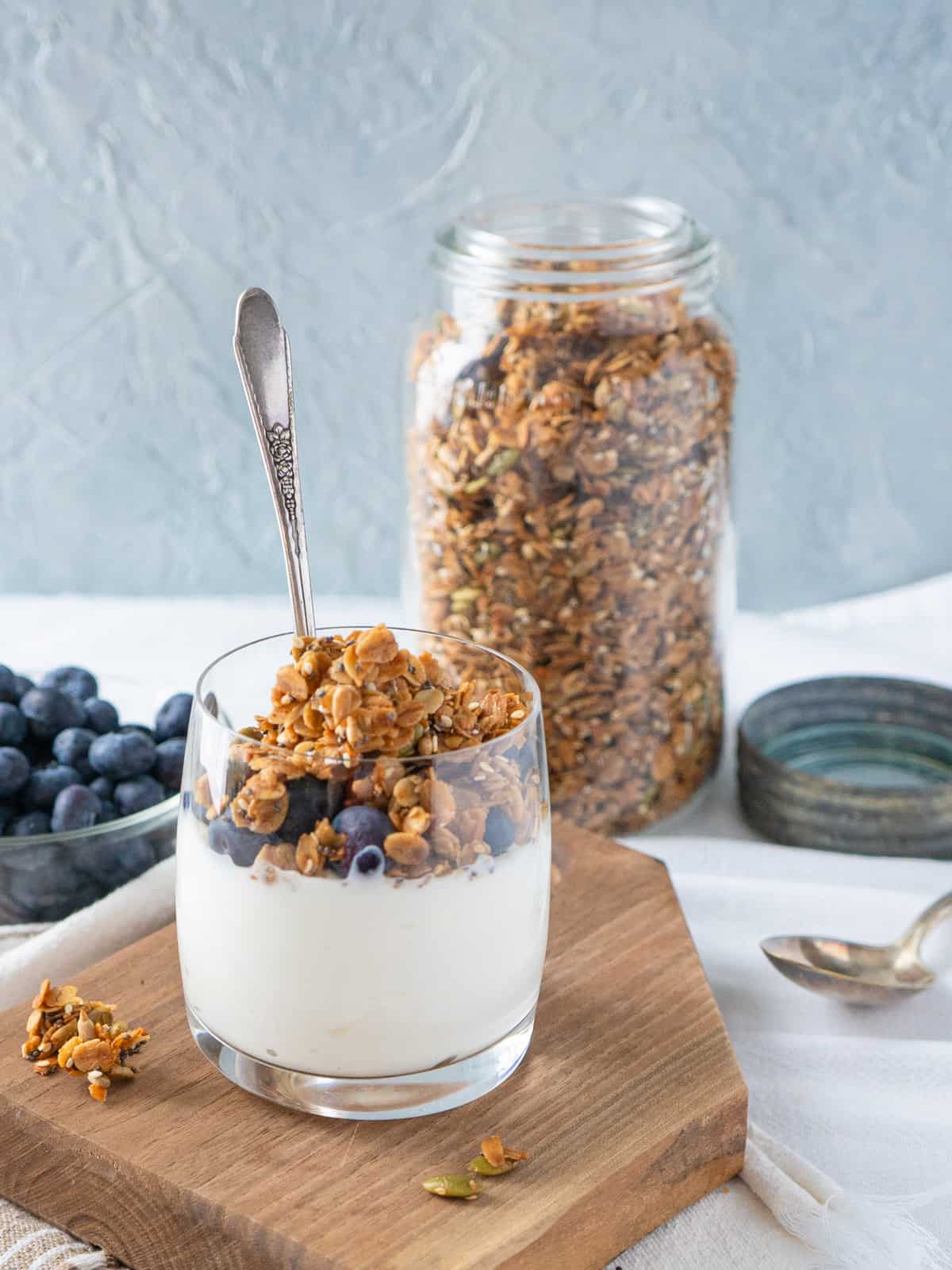 Yogurt in a glass topped with blueberries and granola. Jar of granola and dish of blueberries on the side. 