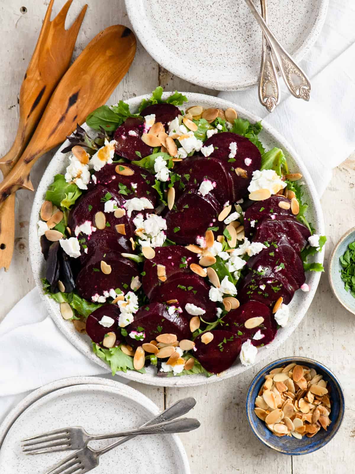 beet salad on a round plate topped with sliced almonds and goat cheese. Plates, serving utensils and a dish of almonds on the side. 