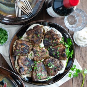 a serving platter of cooked lamb chops on a bed of tahini sauce with a bottle of wine, glasses, plates and fresh parsley on the side.