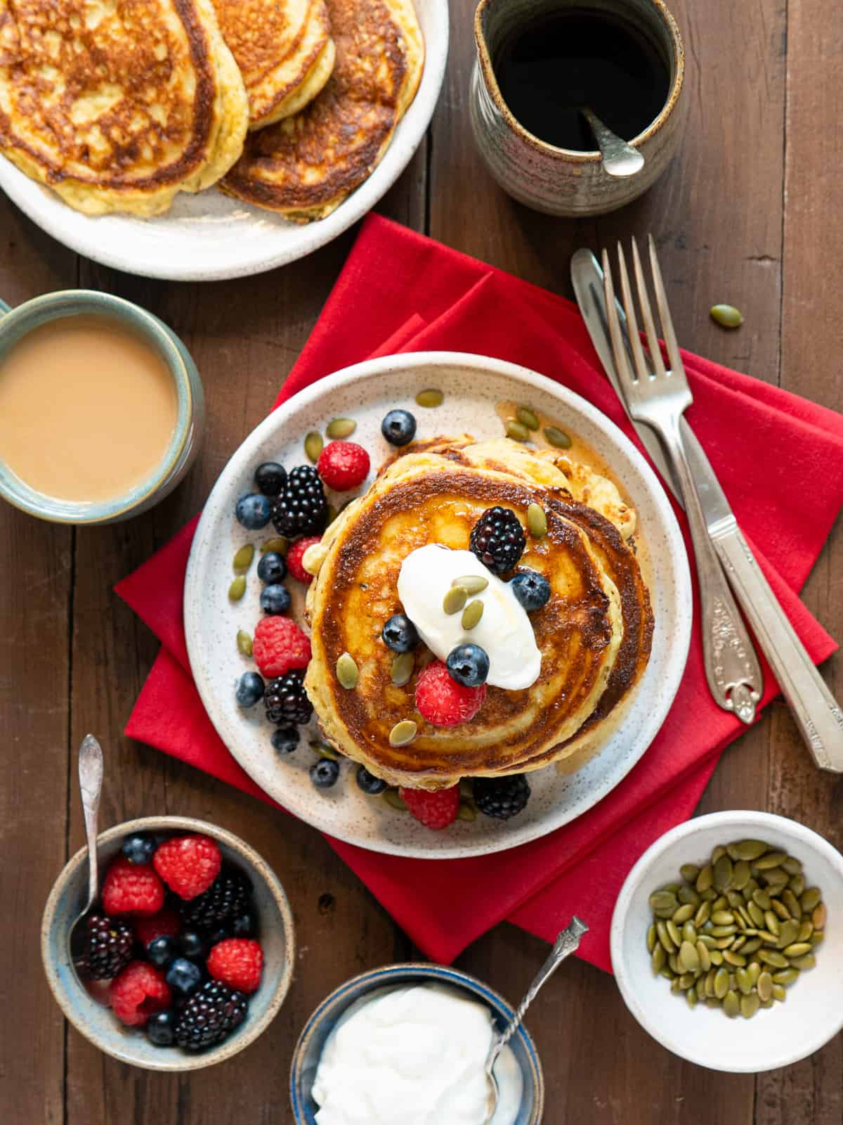 A stack of pancakes on a wood table with a bowls of toppings on the side as well as a cup of coffee and extra plate of pancakes. 