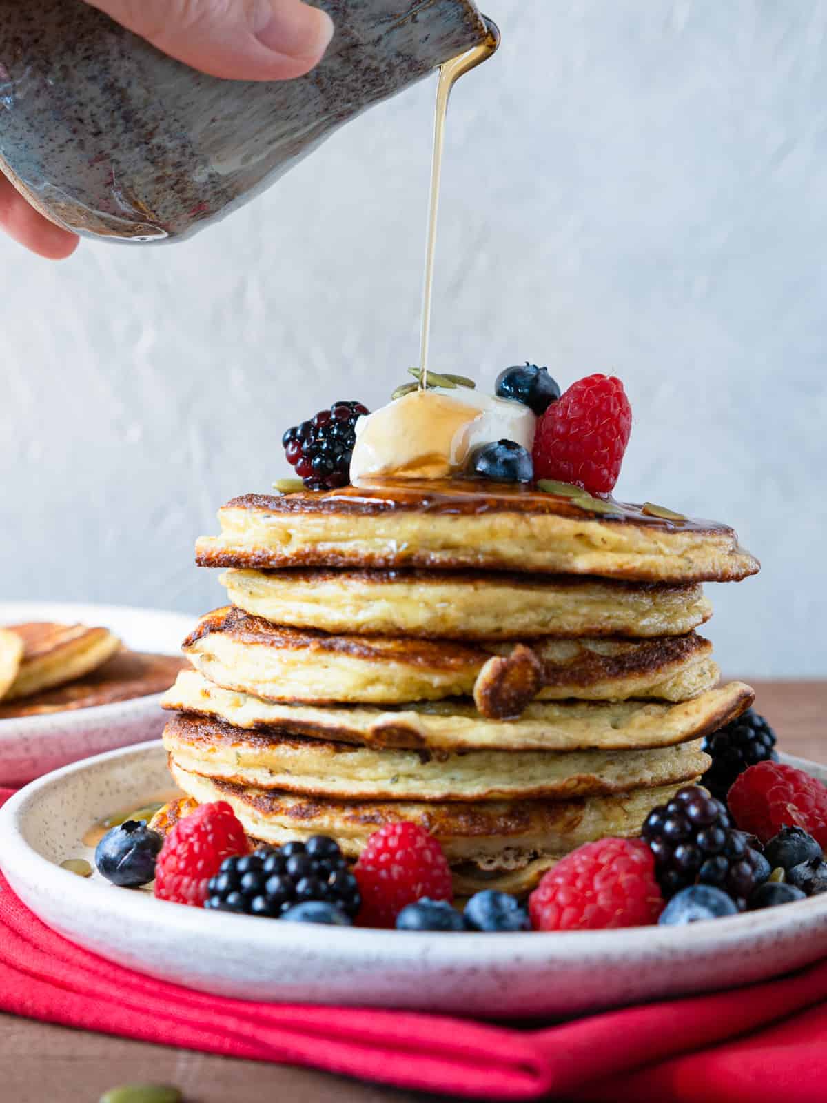 A stack of pancakes on a white plate surrounded by and garnished with fresh berries. On top is a dollop of yogurt and syrup is being poured over the stack. 