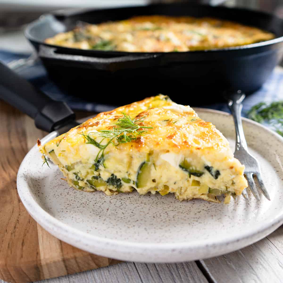 a slice of frittata on a small white plate with a fork on the side. Cast iron pan is in the background. 