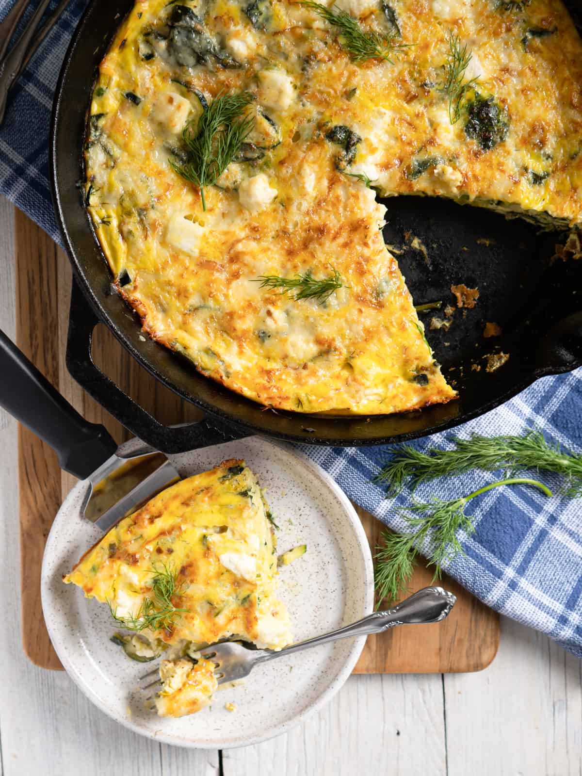 a cast iron pan with baked frittata. One slice has been removed and is on a plate to the side with a bite sized piece on a fork ready to be eaten. 