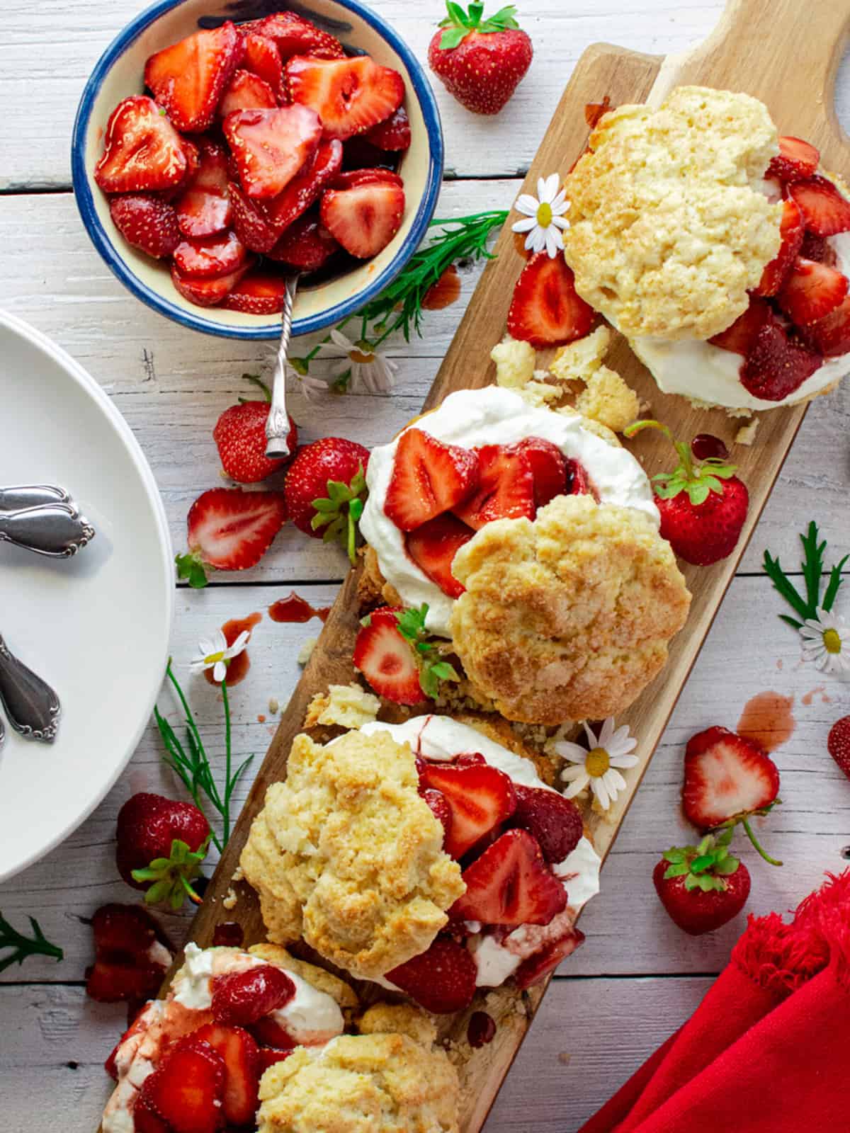 4 cream biscuits filled with whipped cream and strawberries on a wood board. A dish of sliced strawberries is off to the side.