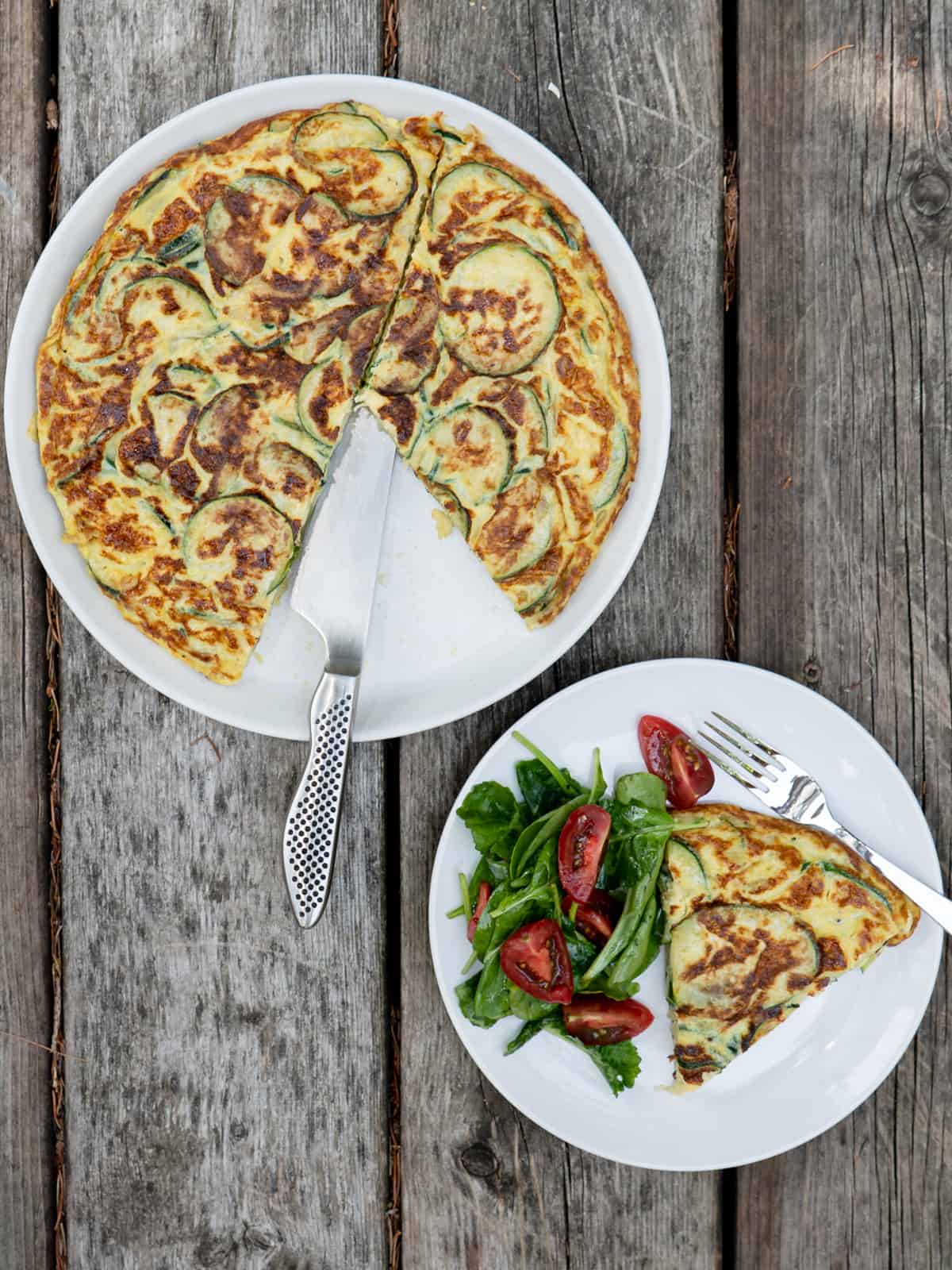 whole omelette on a white plate with a slice removed and on a side plate with a green salad. 