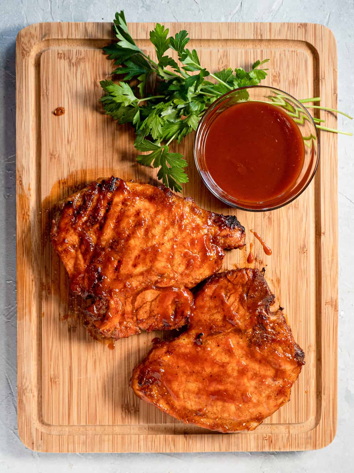 2 bbq'd pork chops on a wood board with a side dish of sauce and fresh parsley. 