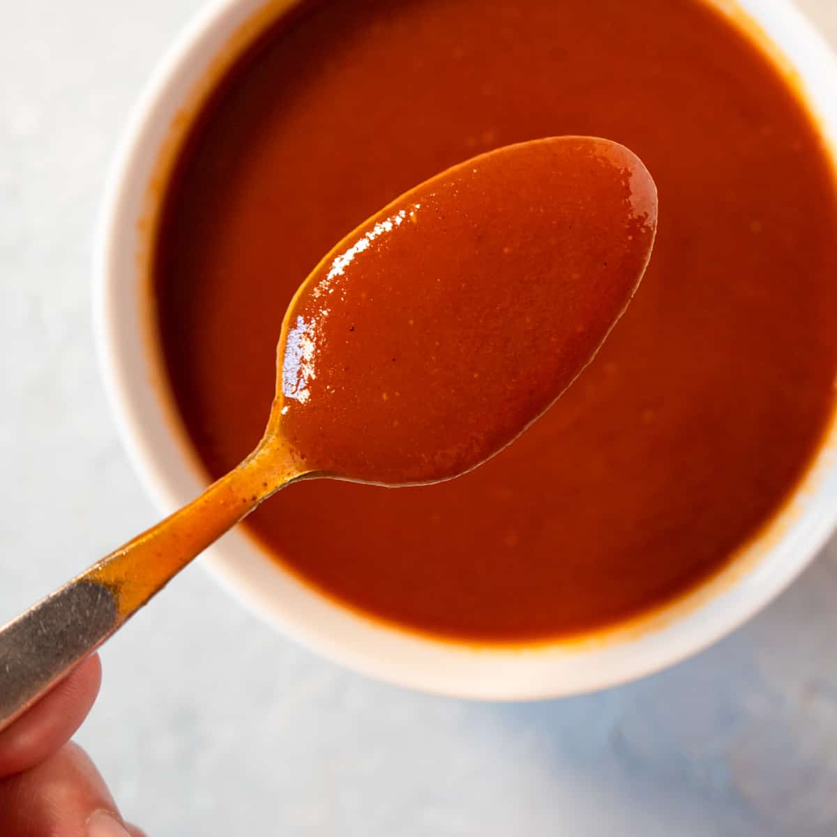 Prepared bbq sauce in a white bowl with a hand holding a spoonful to show how smooth it is.