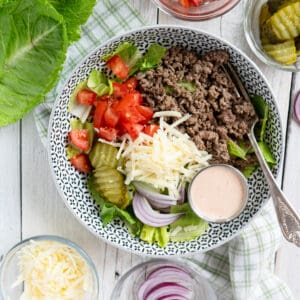 a bowl filled with lettuce, ground beef, tomatoes, onions, pickles and special sauce. Small dishes of toppings are on the side.