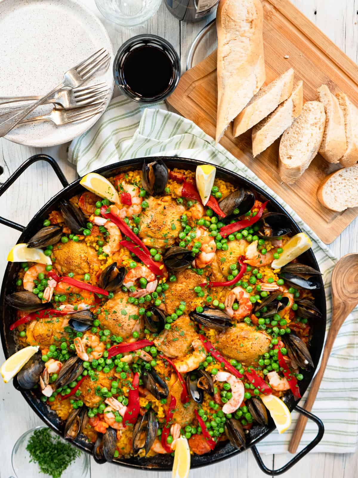 a colorful paella garnished with lemon wedges with a glass of red wine and bread on the side. 