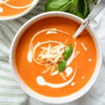 one bowl of roasted tomato and red pepper soup garnished with cheese, cream and basil and a spoon.