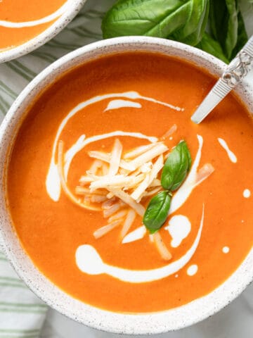 one bowl of roasted tomato and red pepper soup garnished with cheese, cream and basil and a spoon.