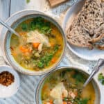 2 bowls of tuscan white bean soup with spoons resting in the bowls. Sliced seedy bread is on the side and soup is garnished with Parmesan cheese.