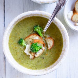 Creamy broccoli soup in a white ceramic bowl with a spoon garnished with croutons and florets.