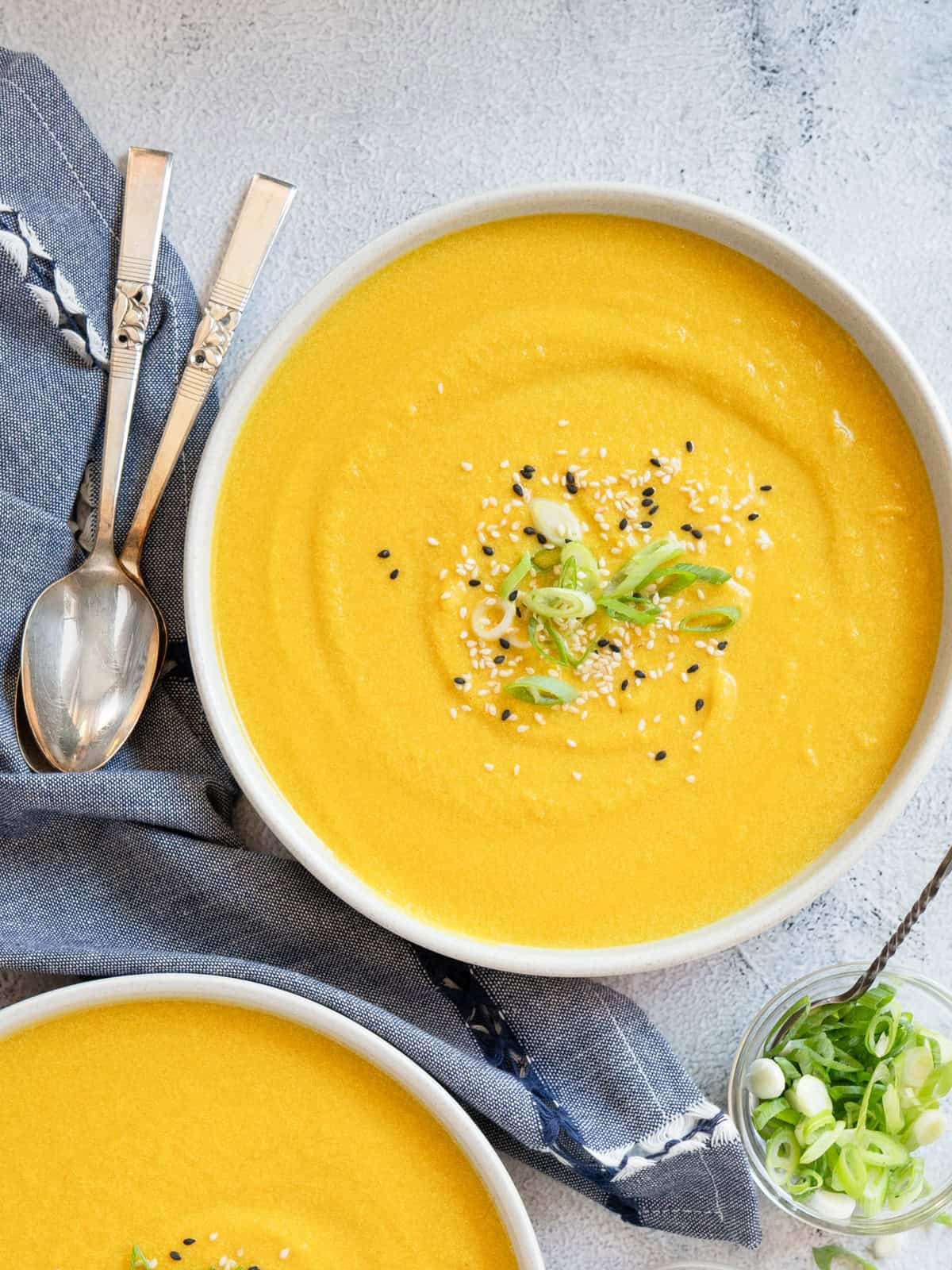 One bowl of soup with another off to the side with spoons on the side as well as a small dish of sesame seeds and another of green onions.