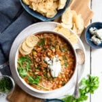 Slow-cooked buffalo chicken chili in a serving bowl topped with sour cream, blue cheese and tortilla chips.
