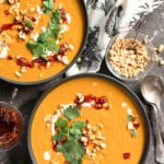 2 bowls of pumpkin soup on a dark background garnished with fresh cilantro leaves, chopped peanuts and chili crisp.