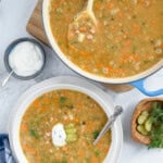 Dill pickle soup in a serving bowl garnished with sour cream. Full pot of soup is in the background with a wooden spoon.