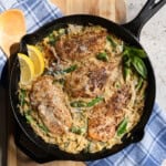 4 chicken cutlets in a bed of creamy orzo with asparagus in a cast iron pan garnished with fresh lemon wedges and fresh basil.