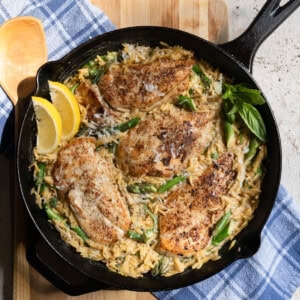 4 chicken cutlets in a bed of creamy orzo with asparagus in a cast iron pan garnished with fresh lemon wedges and fresh basil.