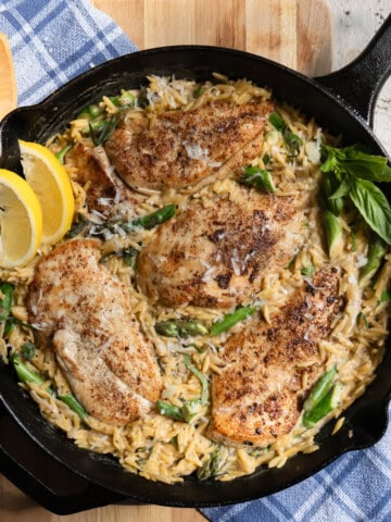 4 chicken cutlets in a bed of creamy orzo with asparagus in a cast iron pan garnished with fresh lemon wedges and fresh basil.