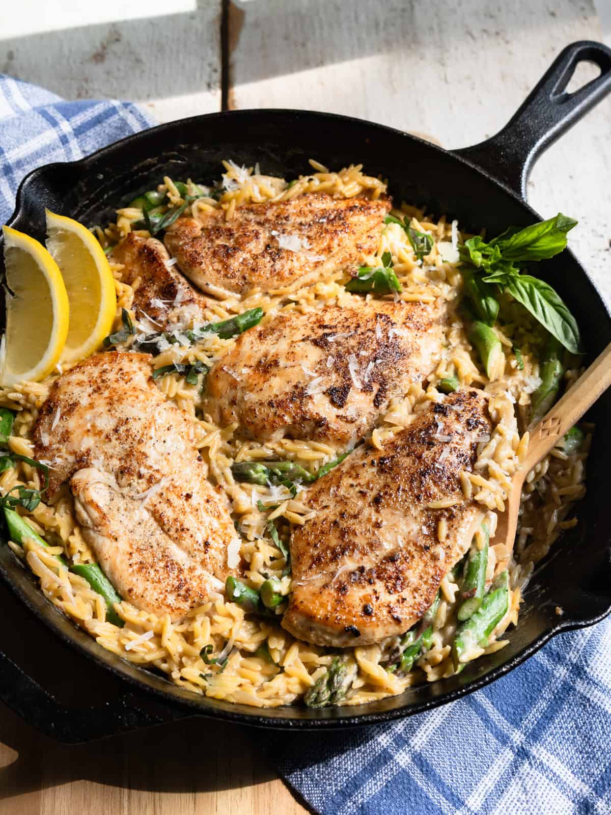 4 chicken cutlets in a bed of creamy orzo with asparagus in a cast iron pan garnished with fresh lemon wedges and fresh basil.