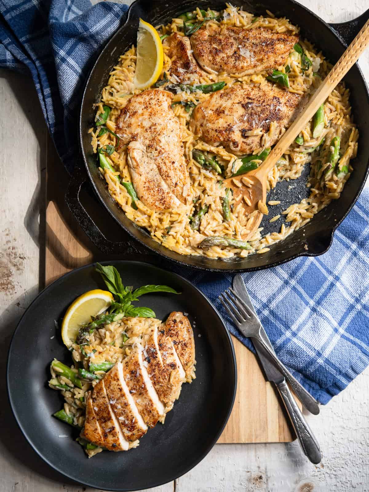 4 chicken cutlets in a bed of creamy orzo with asparagus in a cast iron pan with one serving on the side on a black plate.