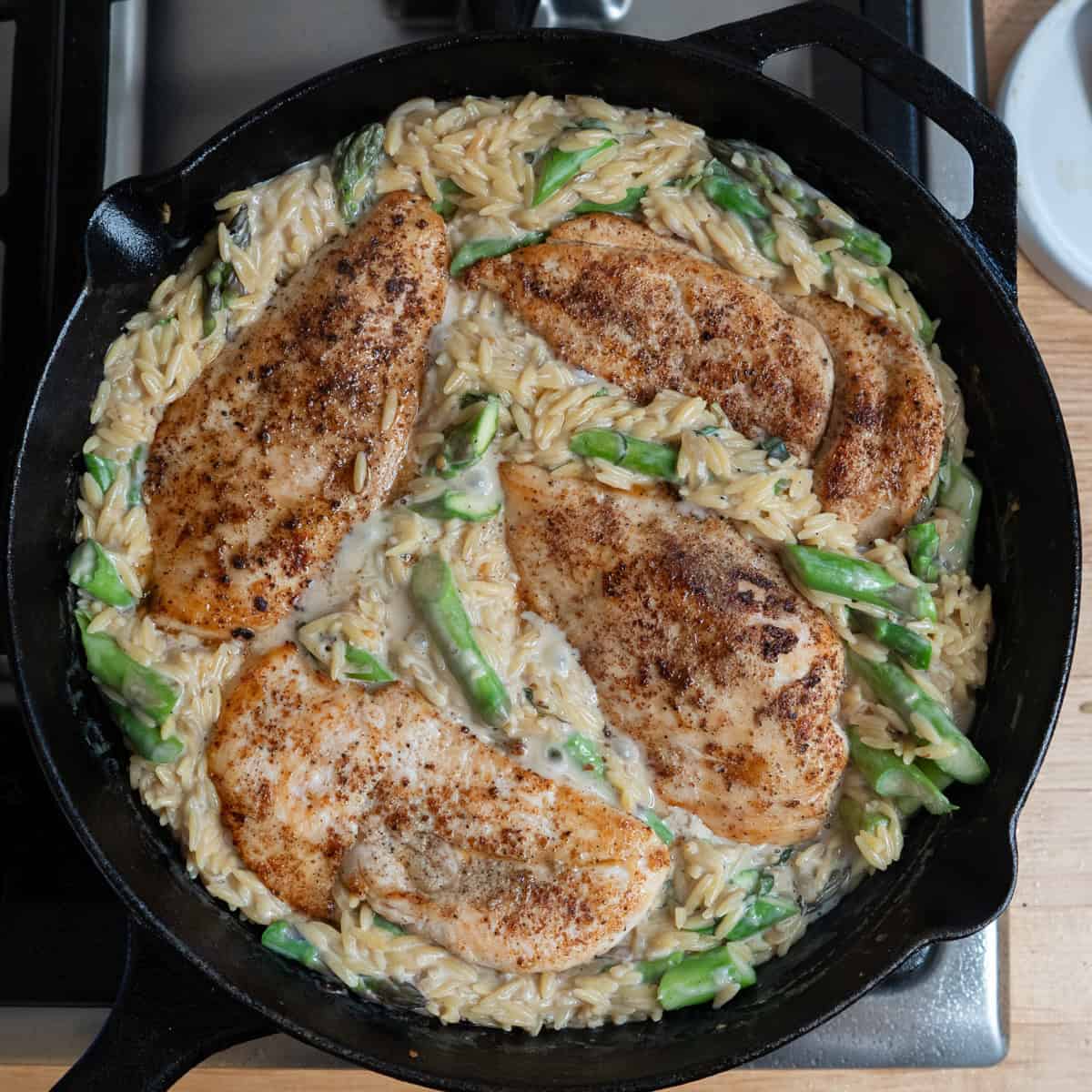 Chicken cutlets in a creamy parmesan orzo with asparagus cooking on the stove in a skillet.