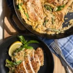 4 chicken cutlets in a bed of creamy orzo with asparagus in a cast iron pan with one serving on the side on a black plate.