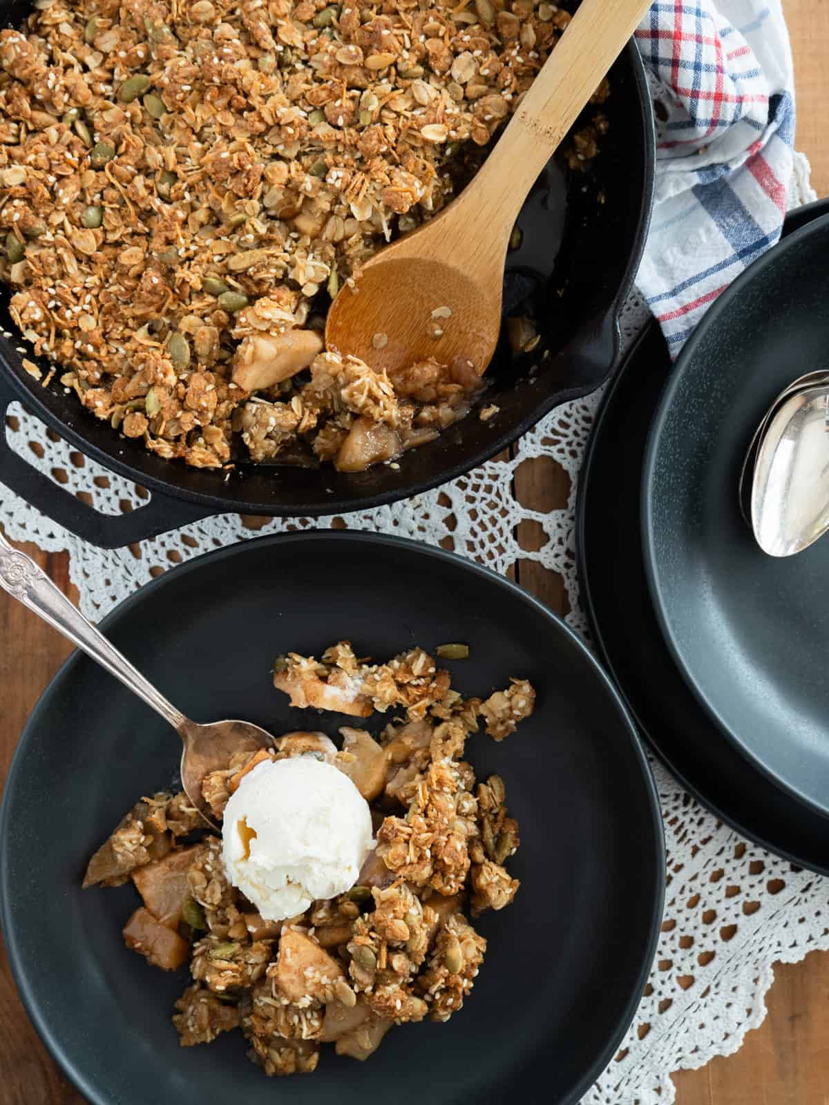 Apple crisp in a cast iron skillet. One serving has been scooped into a shallow bowl and topped with a scoop of vanilla ice cream.