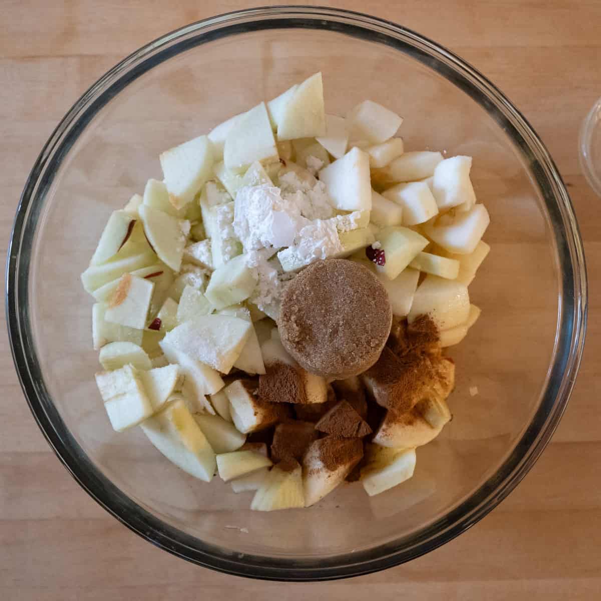 Raw, sliced apples in a glass bowl with Brown sugar, cornstarch, lemon juice, vanilla, cinnamon and salt on top waiting to be mixed together.